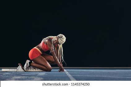 Female Athlete Looking Back At The Starting Block While Taking Position To Start The Race. Side View Of Female Runner Getting Ready At The Start Line On Running Track On A Black Background.