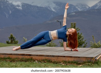 Female athlete in the morning in sportswear, standing barefoot on one side, practicing yoga workout, performs exercises outdoors. flexible strong lady with red hair engaged in sport, yoga - Powered by Shutterstock