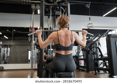 Female athlete performing lat pulldown on cable machine in modern gym from rear view, showing back muscles, focused posture, controlled grip, short hair, sports bra