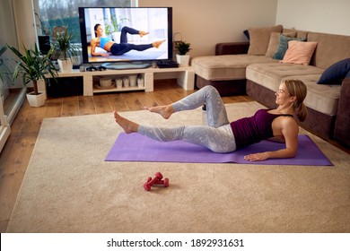 Female Athlete Practicing Her Lower Abs While Following Exercise Class On A TV In The Living Room. 