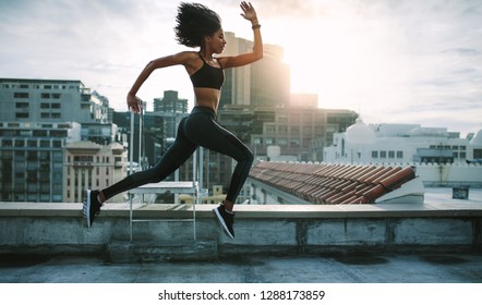 Female athlete running with big strides on rooftop in the morning with sun in the background. Woman in fitness wear training on terrace of a building. - Shutterstock ID 1288173859