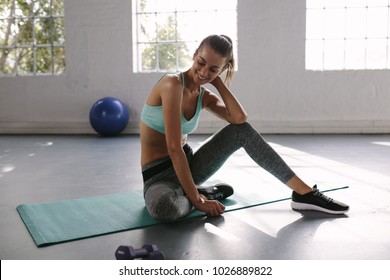 Female athlete sitting on exercise mat at gym and smiling. Fit young woman taking break after her workout at health club.