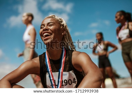 Female athlete smiling after winning a race with other competitors in background. Sportswoman with medal celebrating her victory at stadium.