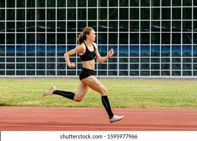 Female Athlete In Spikes Shoes Running Track Stadium In Background Of Glass Facade Building