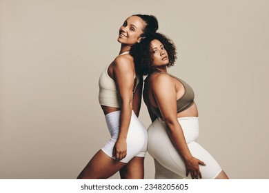 Female athletes of diverse body types stand together back to back in a studio, dressed in workout clothing.Two confident young women showing off their toned bodies and their fitness lifestyle.