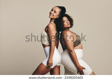 Female athletes of diverse body types stand together back to back in a studio, dressed in workout clothing.Two confident young women showing off their toned bodies and their fitness lifestyle.