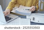 Female auditor in business attire inspecting financial reports with magnifying glass, surrounded by documents and a laptop and calculator. Business audit and taxes concept