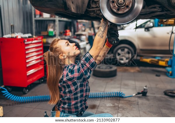 Female auto mechanic. beautiful young red-headed\
girl in working process at auto service station, indoors. Gender\
equality. Work, occupation, car. Pretty girl in work clothes, plaid\
shirt