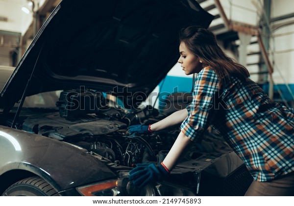Female auto\
mechanic, beautiful young girl wearing plaid shirt in working\
process at auto service station, indoors. Gender equality. Work,\
occupation, car. Pretty girl in work\
clothes