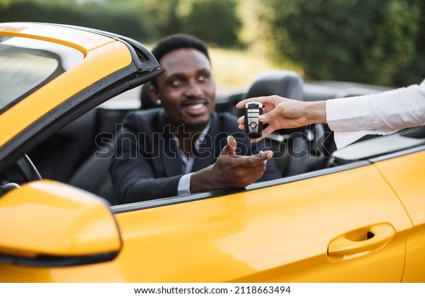 Female auto seller giving keys\
to male customer form his new sport car. Handsome african man in\
suit sitting inside luxury yellow auto. Focus on hands with\
keys.