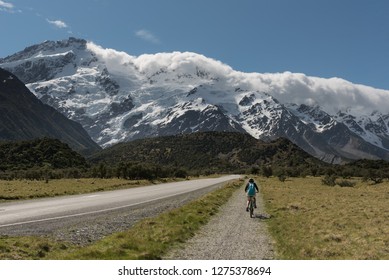 Female, Baby Boomer Cyclist Cycling On A Bike Path Towards The Snow Capped And Glacier Covered Southern Alps, Aoraki/Mount Cook National Park, New Zealand.