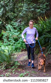 Female Baby Boomer In A Purple Top And Black Running Pants On An Evening Walk With Her Eager Dog On A Forest Trail
