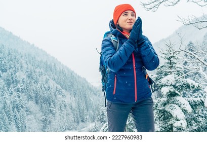Female backpacker with backpack dressed warm down jacket warming palm hands and enjoying snowy mountains landscape while she trekking winter mountain forest route. Active people in nature concept.