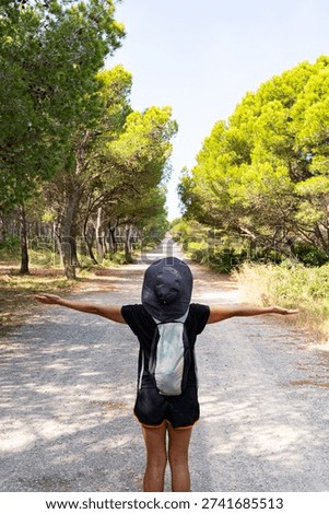 Female backpacker with a hat and backpack spreading arms on a sunny forest path, feeling freedom and wanderlust while hiking