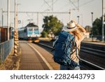 Female backpacker traveling by train alone. Woman with backpack and straw hat standing at railroad station platform and looking at arriving train