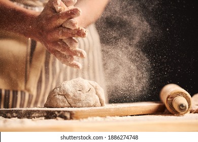 Female Baker Hands Making Dough For Bread With An Apron. Roller And Chopping Board Visible In The Background. Natural Homemade Ingredients. Dark Background, Brown Color Grading.