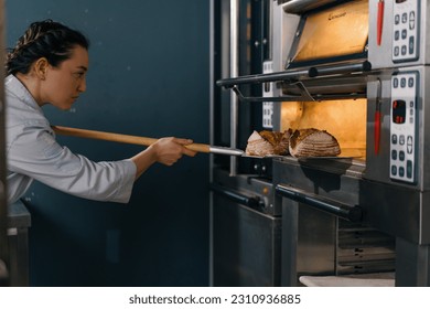female baker takes out freshly baked fresh bread from the oven and puts it on the shelf in kitchen of the bakery Culinary profession - Powered by Shutterstock
