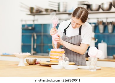 Female bakery chef making delicious sweet cake. Woman applying pink cream from piping bag to sponge layer placed on rotating cake stand in pastry shop kitchen