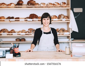 Female Bakery Worker Behind The Counter Ready To Service. Seller Of Zero Waste Shop On Background Of Fresh Organic Homemade Bread On Wooden Shelves In Plastic Free Store. Owner Small Local Business.