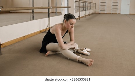 Female ballet dancer finished choreography rehearsal. Tired ballerina in training bodysuit takes off pointe shoes after stretching and gymnastic workout in dance studio. Classic ballet dance school. - Powered by Shutterstock