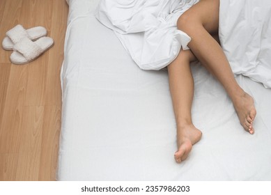 Female bare feet lie on bed under blanket in illuminated bedroom near house slippers on wooden floor in light dwelling lady sleeps sticking out legs on soft place resting - Powered by Shutterstock