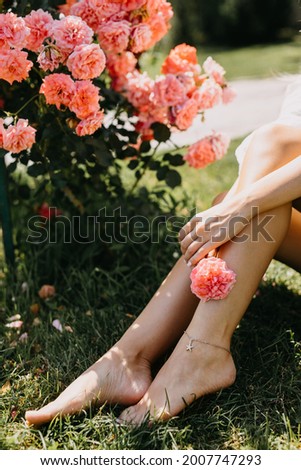 Female barefoot feet with a bracelet on green grass outdoors, next to a bush with pink roses.