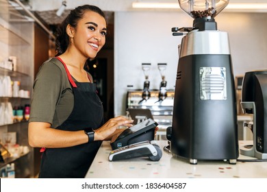 Female barista at counter using cashbox computer in coffee shop. Smiling waitress wearing apron and looking away.