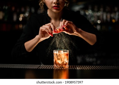 female bartender gently sprinkles orange peel juice on glass with cold alcoholic cocktail on the bar counter