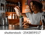 Female bartender holds orange chips with tweezers over a tall glass with a cocktail wrapped in gift paper