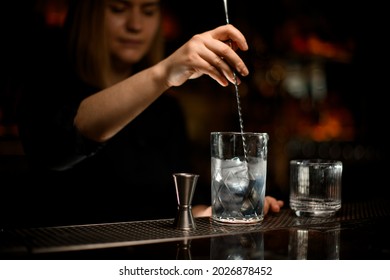 female bartender holds spoon and stirred cold cocktail in crystal mixing cup. Clear old-fashioned glass and steel jigger stand side by side on the bar