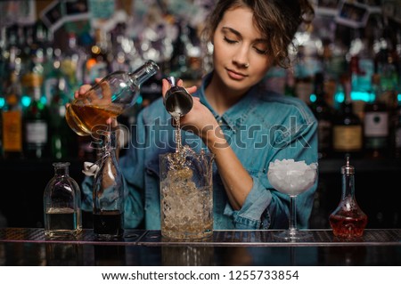 Female bartender pouring to the measuring glass cup with ice cubes an alcoholic drink from steel jigger on the bar counter on the blurred background