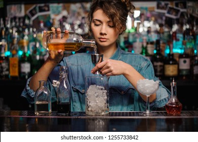 Female bartender pouring to the steel jigger an alcoholic drink on the bar counter on the blurred background