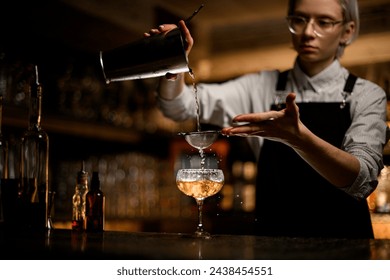 Female bartender pours a clear liquid from a metal beaker into a glass with a sieve - Powered by Shutterstock