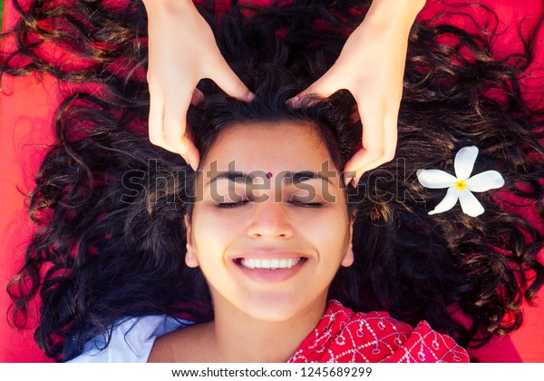 Female beautician doctor with patient make procedure\
to beautiful indian girl.close up portrait of asian woman closed\
eyes and white smile wellness head massaging on beach.Face lifting\
massage in spa