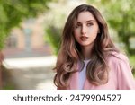 Female beauty. Beautiful girl with gorgeous long wavy hair, dressed in a pink jacket, stands against summer greenery on the city street. Copy space. Summer vacation.