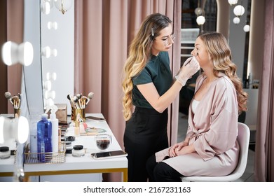 Female beauty specialist in sterile gloves applying foundation on woman face with cosmetic brush. Young woman sitting at dressing table while stylist doing professional makeup in visage studio.