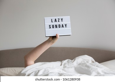Female in bed under the sheets holding up a lazy sunday sign