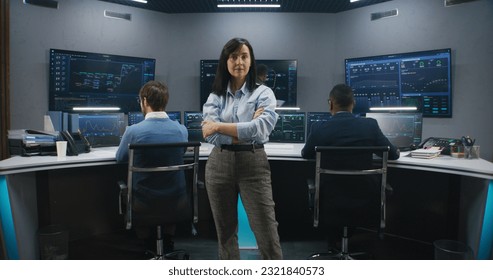 Female big data scientist stands in monitoring room and looks at camera. Software engineers work at computers at background. Multiple big screens with displayed real-time analysis charts. Dolly shot.