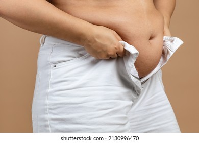 Female with big flabby abdomen trying to zip up white jeans on beige background. Visceral fat. Body positive. Sudden weight gain. Tight little clothes. Need for wardrobe change.