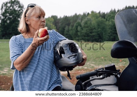 Female biker. Eating apple pretty blonde middle-aged biker near her bike at bus stop during motorcycle rally. Hobbies of strong independent women. Tourism d travel. Adventures. Motorcycle tourism.