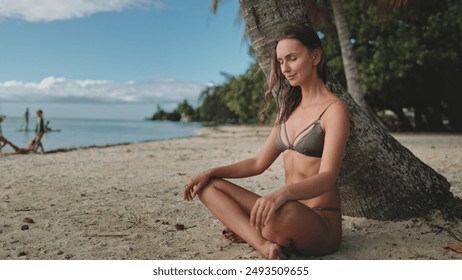 Female in bikini relax under palm tree sitting on sandy beach. She is relaxed, looking on ocean. Long hair slim caucasian brunette enjoy beautiful nature summer landscape. Travel, tourism, holiday - Powered by Shutterstock