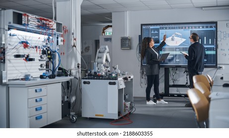 Female Black Developer Looking At Big Screen And Telling Her Ideas To Male Colleague. Robotic Arm Moving In Background. Science And Innovative Technologies On Modern Factory Concept.