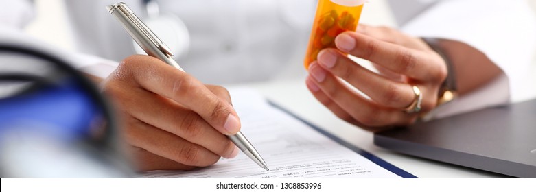 Female black medicine doctor hand hold jar of pills and write prescription to patient at worktable. Panacea and life save, prescribing treatment legal drug store concept. Empty form ready to be used - Shutterstock ID 1308853996