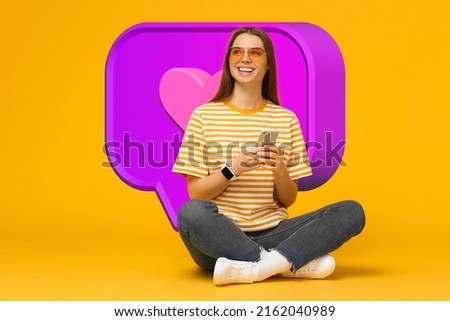 Female blogger in sunglasses wearing smart watch sitting on floor with smartphone happy with likes and comments on her new post, isolated on yellow background with giant speech bubble and heart icon