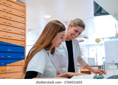 A female blonde pharmacist helps and assists her young aprrentice trainee during studies of medical books