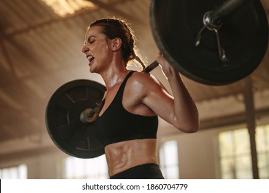 Female bodybuilder doing exercise with heavy weight bar. Fitness woman sweating from squats workout at gym. Female putting effort and screaming while exercising with heavy weights.