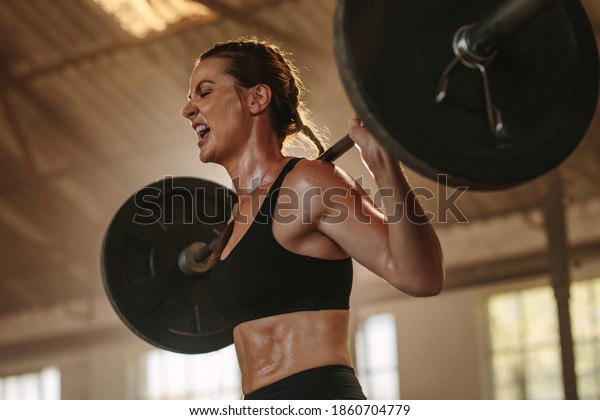 Female\
bodybuilder doing exercise with heavy weight bar. Fitness woman\
sweating from squats workout at gym. Female putting effort and\
screaming while exercising with heavy\
weights.