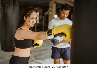 a female boxer standing next to the male boxing coach while punching the heavy bag at the boxing studio - Powered by Shutterstock