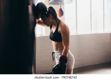 Female boxer taking a break from her practice. Woman boxer leaning forward to rest on a huge punching bag at a boxing studio. - Powered by Shutterstock