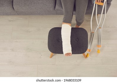 Female broken injured leg relaxing on stool in cast and metal crutches nearby at home over flooring, top view. Trauma, injury, recovery, rehabilitation concept